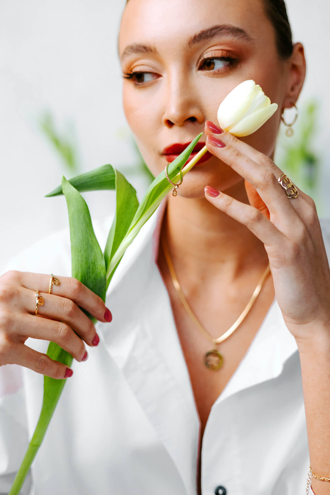 Kobieta w białej koszuli trzyma zielony liść, prezentując złote pierścionki z drobnymi zawieszkami, złoty naszyjnik z okrągłą przywieszką oraz złotą bransoletkę. Na uchu widoczny jest także złoty kolczyk-koło z zawieszką. Biżuteria podkreśla delikatność i elegancję stylizacji.
