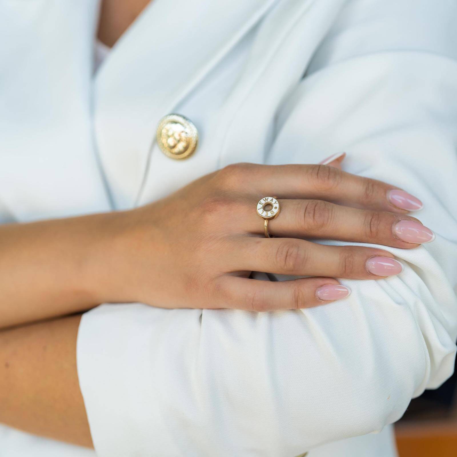 Zbliżenie na dłoń kobiety w białym żakiecie, na której widoczny jest złoty pierścionek z okrągłym, dekoracyjnym elementem. Guzik żakietu również jest złoty, co podkreśla spójność i luksusowy charakter stylizacji.