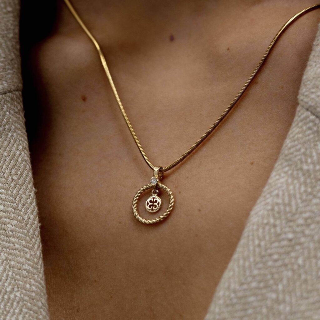 Złoty wisiorek na łańcuszku na dekolcie kobiety ubranej w beżową marynarkę.