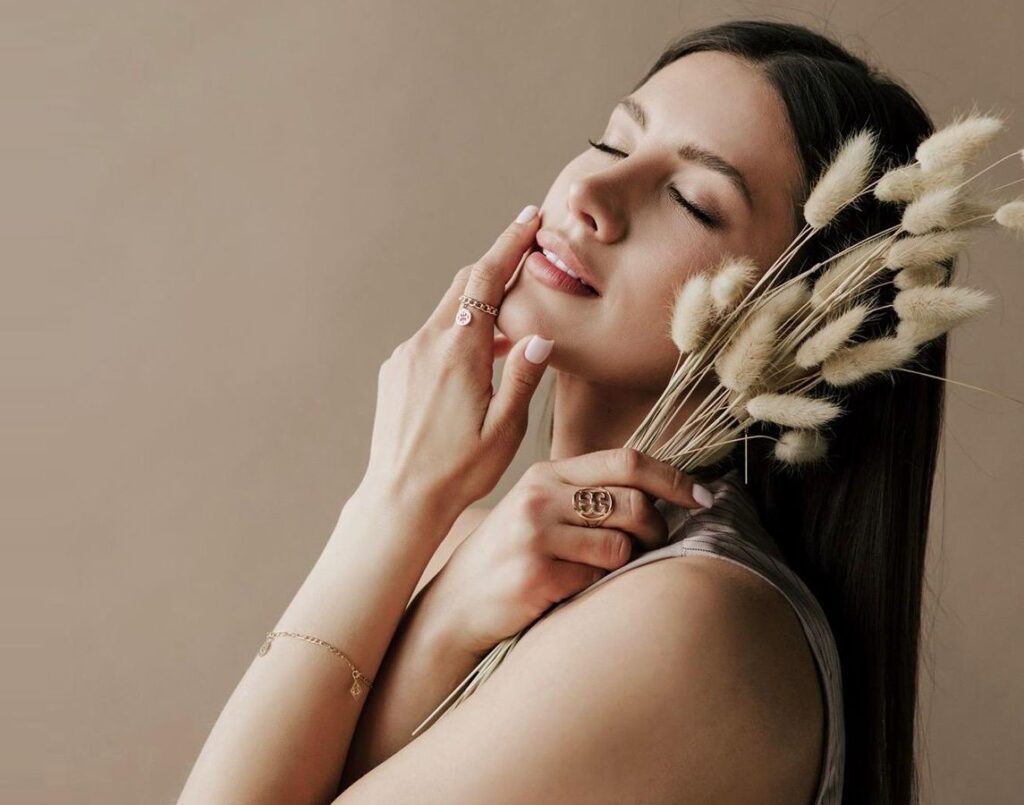 Kobieta z dłońmi przy twarzy na których ma złote pierścionki i trzymająca kłosy traw