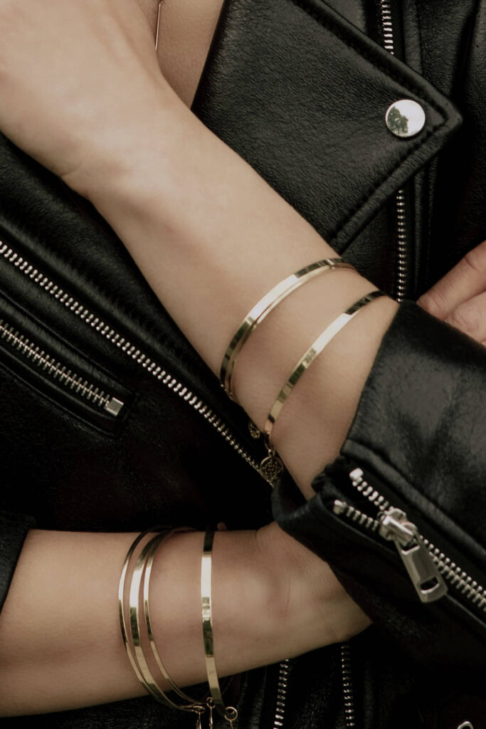 Złote bransoletki twarde z wisiorkami na obu rękach modelki ubranej w czarną skórzaną kurtkę
