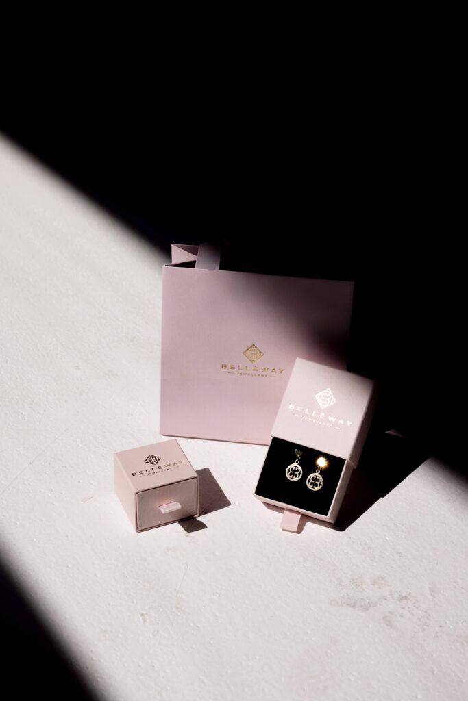 Zlote koczyki wiszące umieszczone w ozdobnym różowym pudełeczku. Obok drugie  różowe pudełeczko i papierowa torebla z nazwą Belleway na białym tle