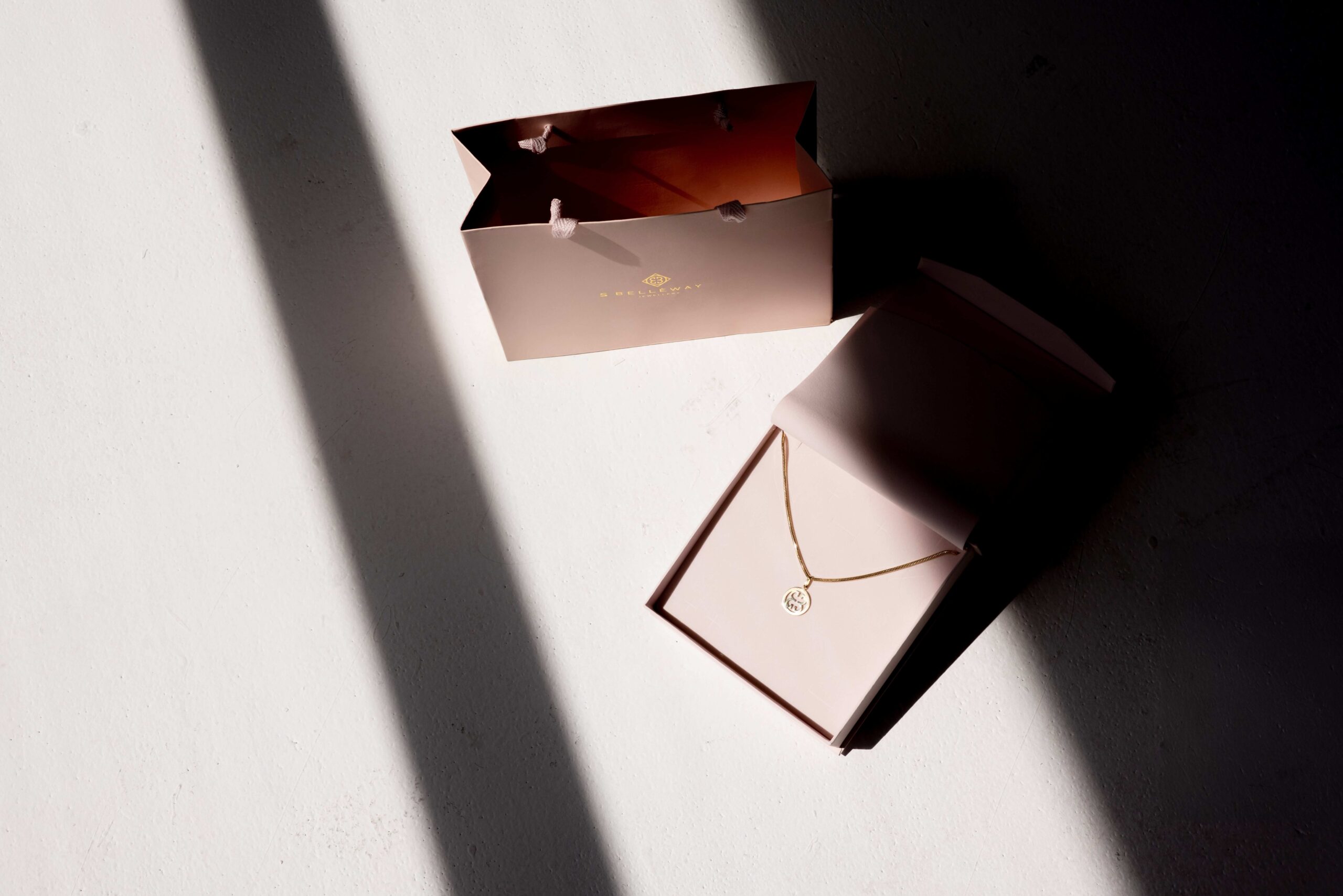 Zloty łańcuszek z zawieszką Belleway W różowym pudełku .obok stoi róowa papierowa torebka marki na białym tle.