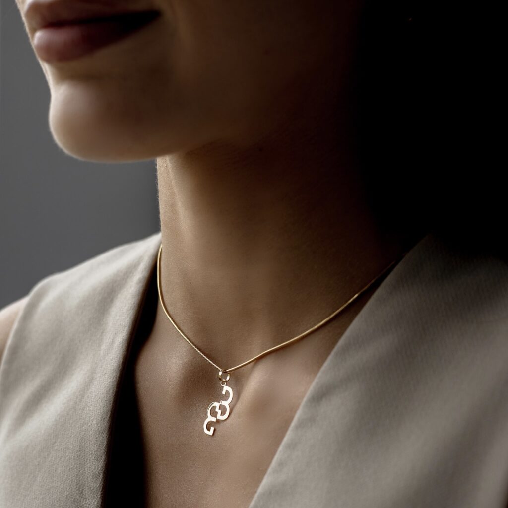 Złota zawieszka na łańcuszku na szyi kobiety ubranej w beżową kamizelkę.