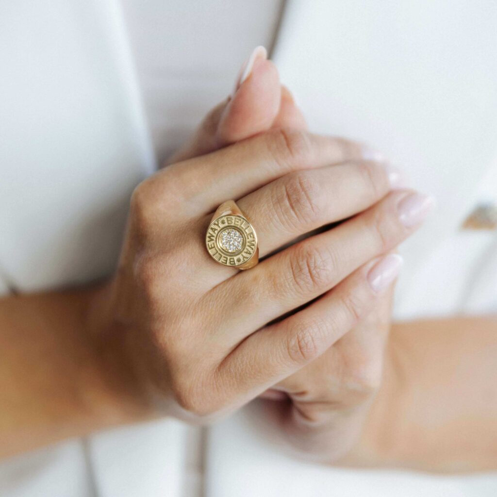 Złoty pierścionek w formie sygnetu wysadzany kamieniami na splecionych dłoniach i na tle białej marynarki
